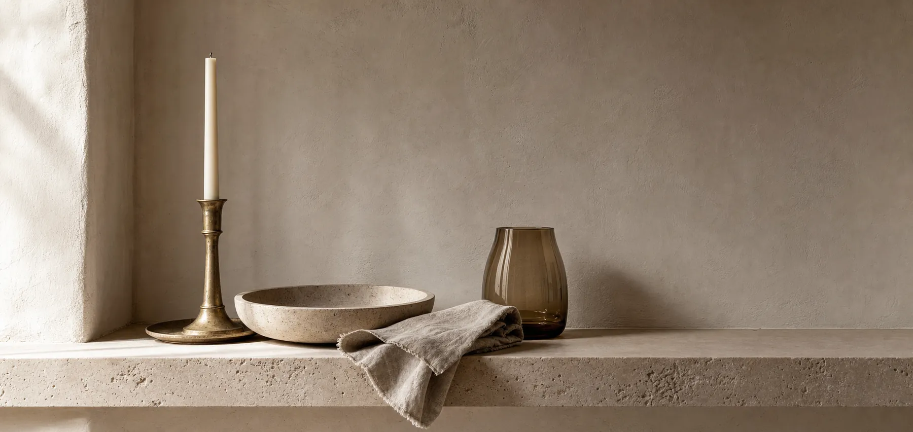 A considered arrangement of a brass candlestick, travertine bowl, linen and amber glass.