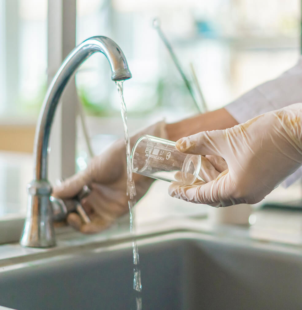 A person wearing latex gloves and a lab coat while holding a glass vial turns on a faucet.