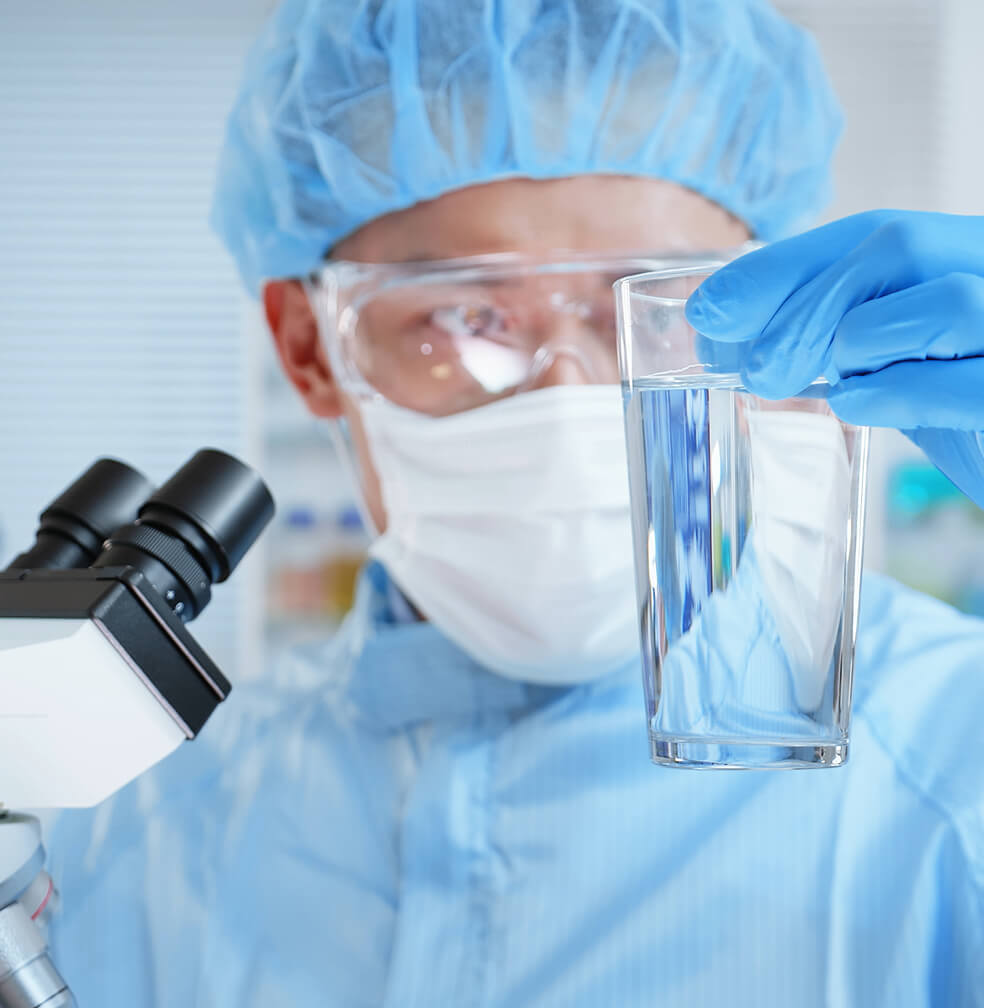 A scientist wearing protective layers inspects a glass of water.