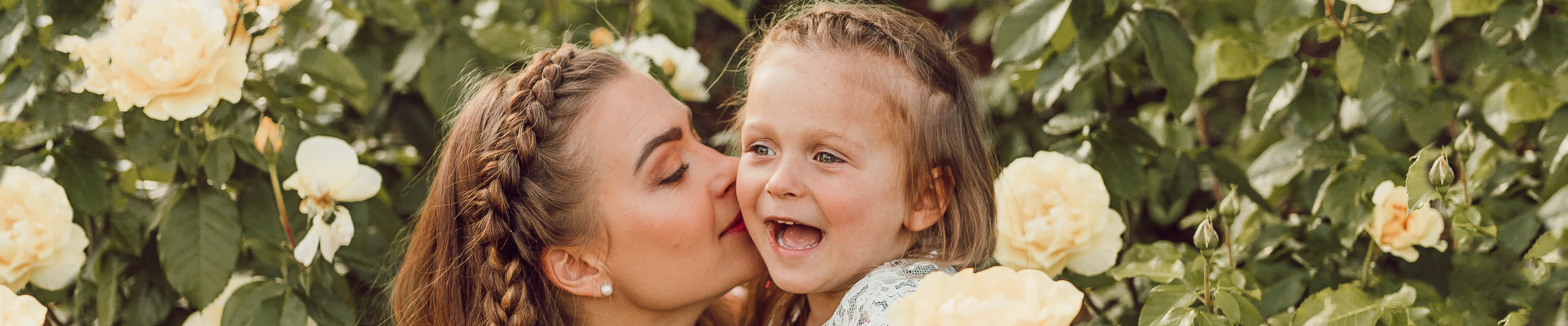 mother kisses the cheek of young daughter in garden full of white flowers