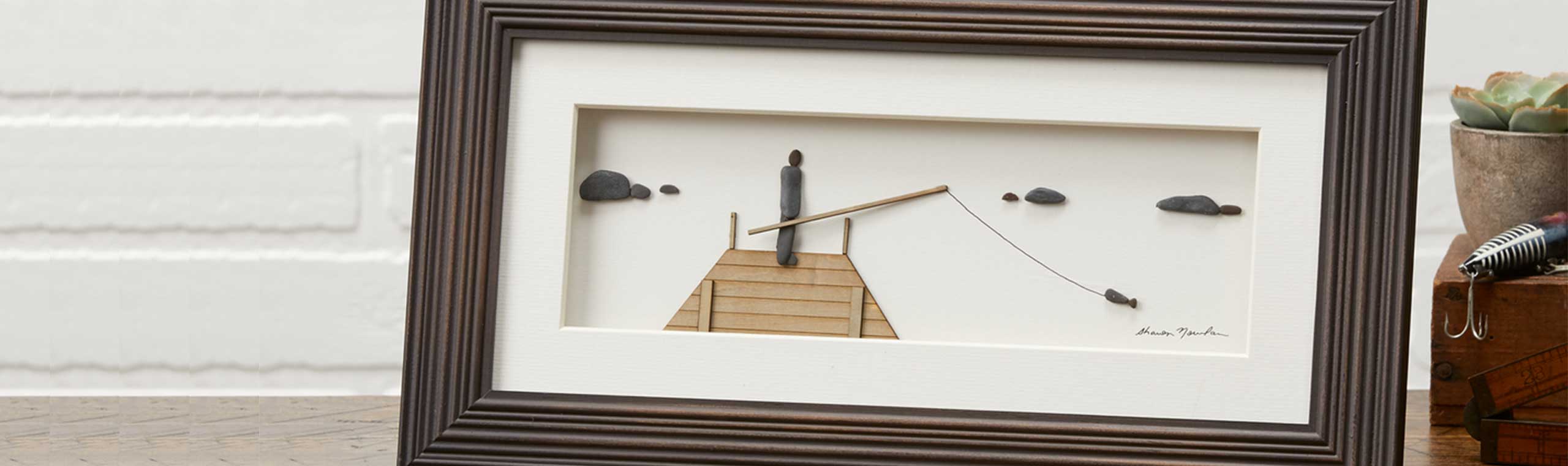 Wood-framed pebble art of man fishing off a dock
