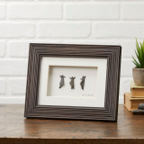 Brown grooved frame with white mat board and background. Three pebbled people are dancing. Frame sits on a wooden shelf. White brick background