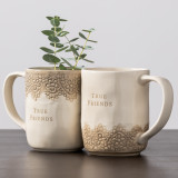 A set of two cream coffee mugs, each with a brown lace pattern and reads "true friends". Placed on a wooden table.
