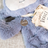 Close up detail view of a blue gray plush cute monster blankie with a "Let's Be Brave" packaging tag tied about its neck.