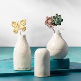 A teal tabletop, topped with three cream vases, each with a varying shape and simple message, Two filled with ceramic and wire flowers.