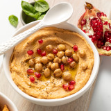 A close up image of a white ceramic bowl filled with hummus. Placed on a wooden tray with fresh herbs and a matching spoon.