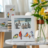 A close up image of a collection of Dean Crouser items placed on wooden and marble side tables. Two plaques with watercolor painted birds and messages, and one topped with a picture frame with watercolor painted birds and a vase of yellow flowers.