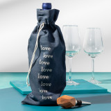 A teal tabletop, topped with two empty wine glasses, a wooden cork, and a bottle of wine wrapped in a blue wine bag that reads "love" several times in white font.