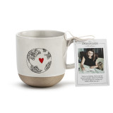 A cream stoneware handled mug with a tan textured base and an image of a scribbled earth that has a red heart inside and a product tag attached to the handle.