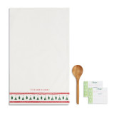 A white tea towel with small red pom pom stripes, and a printed green Christmas tree pattern at the bottom. Placed to the left of a set of recipe cards and a wooden spoon.