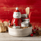 A woven fabric basket filled with a variety of red and white holiday kitchen products, including a Santa cookie jar, a hot pad and spatula set, Santa and snowman toothpick holders and snack bowls. The basket is displayed on a wood surface in front of a red wood background.