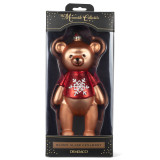 A blown glass ornament of a light brown little bear, wearing a red shirt with a large white snowflake. The ornament is hanging on a red ribbon, displayed in a packaging box.