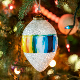 An onion shaped hanging ornament with an image of vertical paint stripes in shades of blue and orange around the middle and silver glitter around the top and bottom inspired by artwork from ArtLifting artist Charlie French, displayed hanging in a lit Christmas tree.