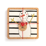 A square ceramic trivet with horizontal black lines and a snowman sitting in a cork base, displayed with a packaging tag.