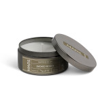 A round dark metal travel tin candle with the lid off and propped on the side for the Smoked Woods scent.