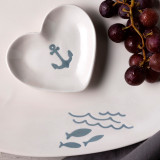 Close up of a white ceramic platter that has waves and blue fish. On the platter is a heart shaped white trinket dish with a blue anchor.
