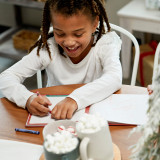 A smiling young girl drawing with crayons in a Santa Kindness journal at a holiday table. A smiling young girl drawing with crayons in a Santa Kindness journal at a holiday table.