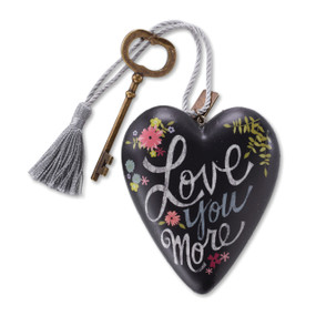 A black heart shaped sculpture that says "Love You More" with a silver tassel and gold key attached.