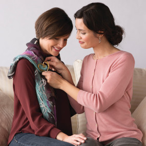 Two women sitting down - one is putting gold heart pendant on the other
