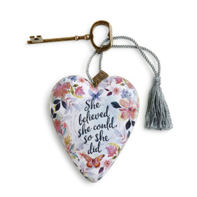 A white floral heart shaped sculpture that says "She believed she could, so she did" with a silver tassel and metal key attached.