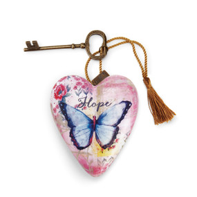 A light pink heart shaped sculpture with a blue butterfly and the word "Hope" with a gold tassel and metal key attached.