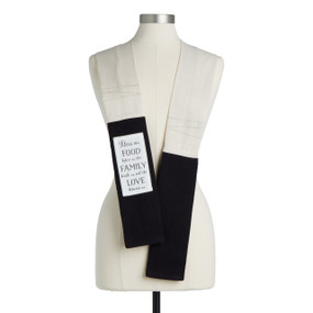 A tan fabric kitchen boa with black towel ends and a white fabric patch that says "Bless this Food before us, the Family beside us, and the Love between us" displayed on a mannequin.