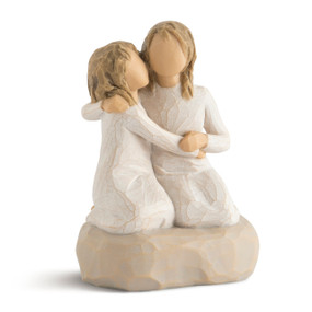 Front view of two female figures wearing cream dresses with arms around each other, kneeling on short beige colored pedestal