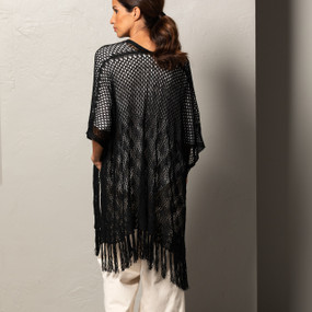 A close up image of the backside of a woman wearing ivory capri pants, a white blouse, and a crochet black duster. Standing in front of a textured gray wall.