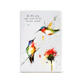 A white ceramic plaque with two painted hummingbirds flying above a red flower, and be the joy you wish to see in the world." in black cursive font."