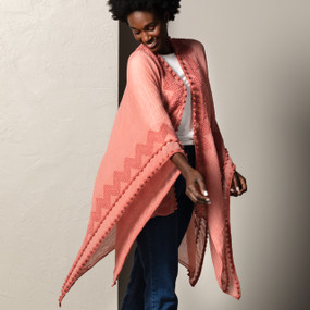A close up image of a woman standing against a textured gray wall, wearing denim jeans, a white top, and a coral duster with a single chevron line and pom pom stripes along the bottom.