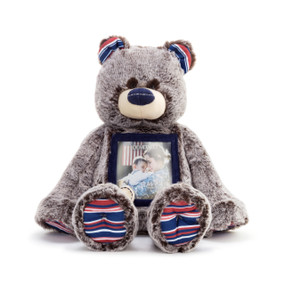 A front facing view of a soft gray and brown plush Here to Hug" bear with red, white, and blue striped paws and ears, a blue nose, a blue photo frame on the stomach, filled with a family photo, and an ivory heart shaped patch that reads "here to hug" in blue stitching."