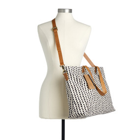 A white tote bag with black triangles that has a leather shoulder strap and handles displayed on a mannequin.