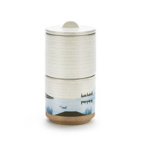 A three piece round ceramic stacked container in cream. The bottom container has a dock and lake scene in blue colors.