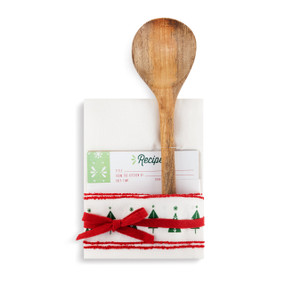 A white tea towel with small red pom pom stripes, and a printed green Christmas tree pattern at the bottom. Tied with a red bow, and filled with a set of recipe cards and a wooden spoon.