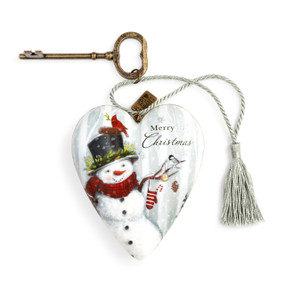 A white Art Heart with an image of a decorated snowman, topped with two birds, in front of a forest scene. with a bronze key and silver tassel.