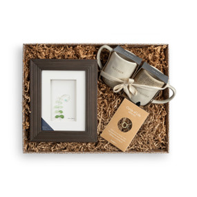 A brown gift box filled with a Circle of Life Gold/Green necklace, mother and daughter coffee mugs, and a brown frame enclosing pebbles depicting a leafy stem.