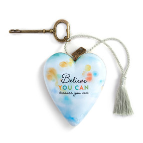 A heart shaped sculpture with a gold key and silver tassel featuring artwork created by ArtLifting artist Eve Hennessa. The image has pale pastel shapes with "Believe You Can because you can" on a light blue background.