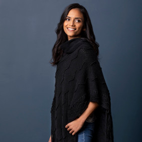 A smiling woman wearing a black textured black cowl neck poncho in front of a dark blue background.