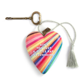 A heart shaped sculpture with a gold key and silver tassel featuring artwork created by ArtLifting artist Jeff Roysdon. The image is bright lines of color and says "Ain't You Just A Little Ray of Sunshine".