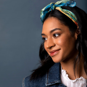 A smiling woman wearing a blue and green scarf tied as a headband. The scarf is inspired by artwork created by ArtLifting artist Dale Wayne.