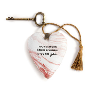 A heart shaped sculpture with a gold key and gold tassel with a pink and white marble background that says "You're Strong You're Beautiful & You Are You".