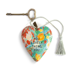 A heart shaped sculpture with a gold key and silver tassel featuring artwork created by ArtLifting artist Alicia Sterling Beach of a bright colorful floral scene on a red background that says "You Lovely Thing, You".