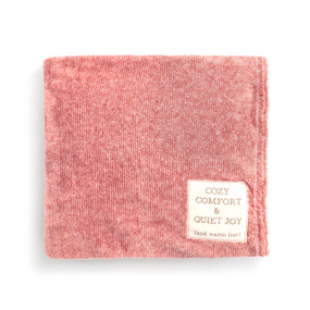 A light red blanket folded and has a fabric tag saying "Cozy Comfort & Quiet Joy (and warm feet)".