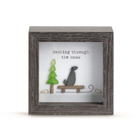 A square dark wood shadow box with a dog on a sled next to a green tree and says "dashing through the snow" made from glass pebbles.