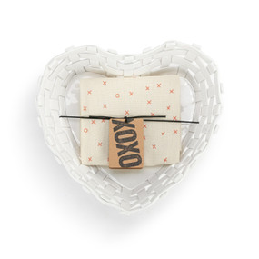 Top down view with the packaging tag for a white ceramic woven heart shaped basket with a cream towel rolled inside.