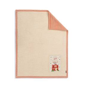 A light brown blanket with a red and brown striped back and a fabric patch in the bottom right corner with a barn on it.