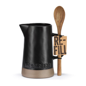 A black ceramic handled pitcher with a brown textured base that says "Refresh" displayed next to a long wood spoon, displayed with a product tag attached.