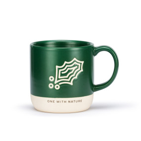 A green ceramic mug with a holly leaf shaped cream meditation path on the mug and a white textured base that says "One With Nature".