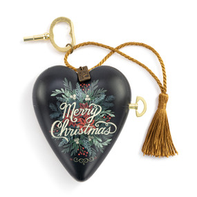 A black heart shaped musical sculpture with a holiday floral pattern and the saying "Merry Christmas". The heart has a gold tassel and gold key attached.