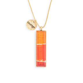 An adjustable gold chain necklace with a vertical gold charm with orange pattern artwork inspired by ArtLifting artist Christina Constantine. The necklace also has a small round gold token that says "courage".